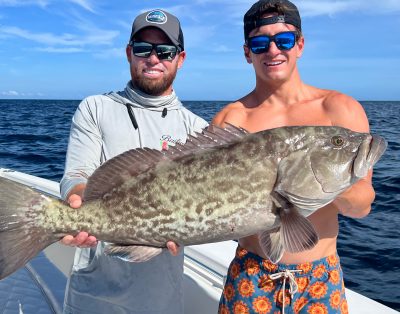 Two friends proudly holding a grouper they caught during a fishing charter in Melbourne, Florida, with the ocean and a blue sky in the background.
