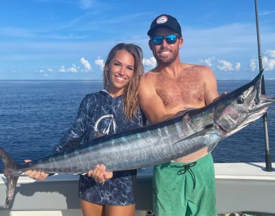 A couple happily holding a wahoo fish caught during a fishing charter in Fort Lauderdale, Florida, with the ocean and a clear sky in the background.