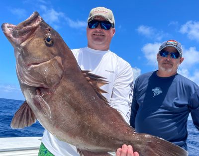 Two friends proudly displaying a massive Warsaw grouper they caught during their fishing charter trip.
