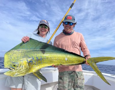 A couple proudly displaying a massive mahi mahi fish caught during a fishing charter in Key West, Florida, with the ocean and a clear sky in the background.