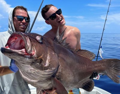 Two friends proudly holding a massive Warsaw grouper they caught during a fishing charter trip.
