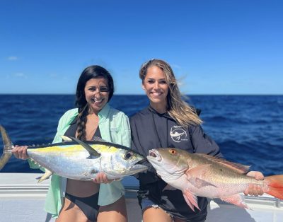 Two women proudly displaying a mutton snapper and tuna caught during a fishing charter on the ocean.