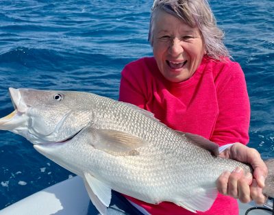 An older woman smiling and holding a margate fish while on a fishing charter, with the ocean in the background.