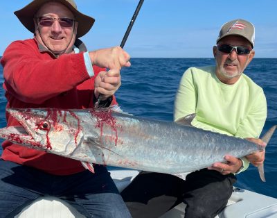 Two older men smiling and holding a big kingfish while on a fishing charter, with the ocean in the background.