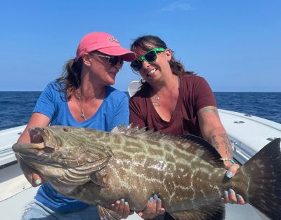 Two best friends, women, showcasing a black grouper caught during a deep-sea fishing trip with their husbands.