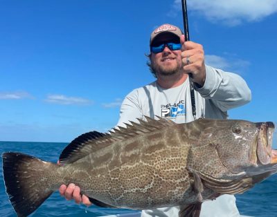 A man smiling for the camera while holding a big black grouper during a fishing charter with friends.