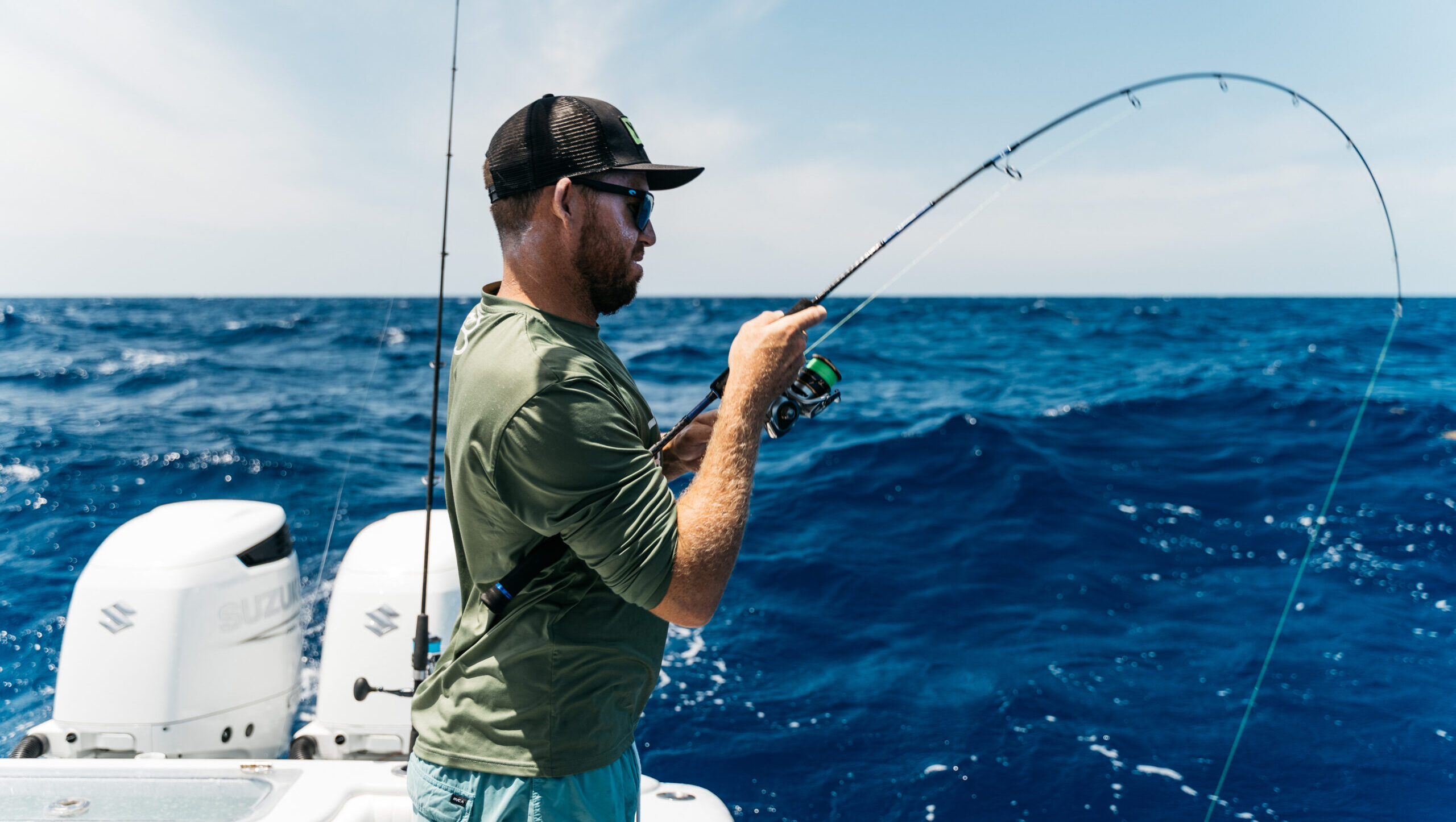 Customer joyfully reeling in a fish surrounded by fishing equipment while on a charter, with a stunning ocean backdrop and the sun shining in the background.