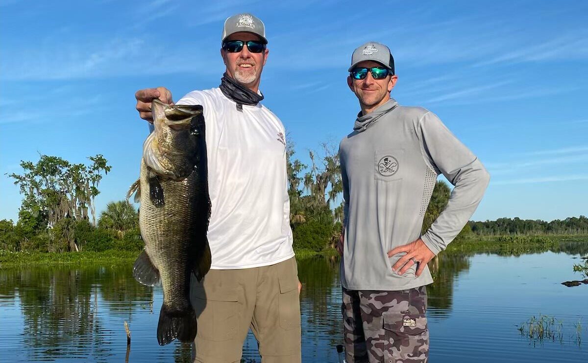 A captain and customer on a freshwater fishing charter in Florida, holding a large mouth bass they caught. The background showcases the serene freshwater environment, enhancing the experience of their successful fishing trip.