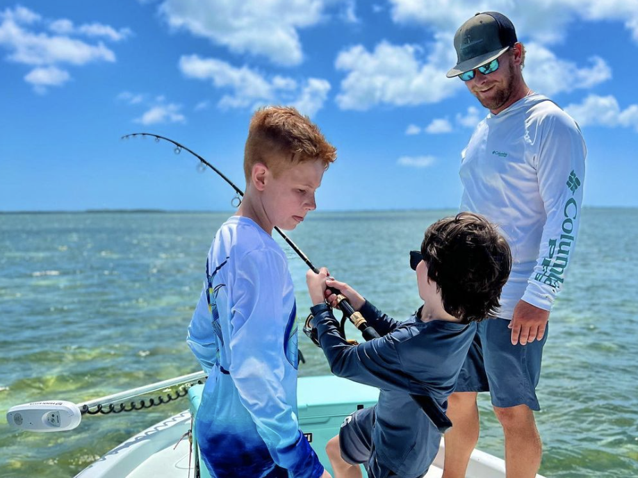 Image of two boys reeling in a fish on a fishing charter boat, with the captain providing guidance and supervision. The scene captures the excitement of learning to fish and the expertise of the captain, creating a memorable fishing experience for the young anglers.