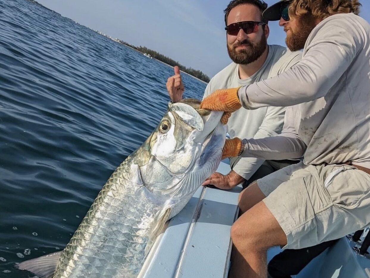 An angler proudly holds up a large tarpon, showcasing a successful fishing catch aboard a fishing charter. The tarpon's silvery scales glisten in the sunlight, and the background features a scenic view of the open sea with a clear blue sky and calm waters.