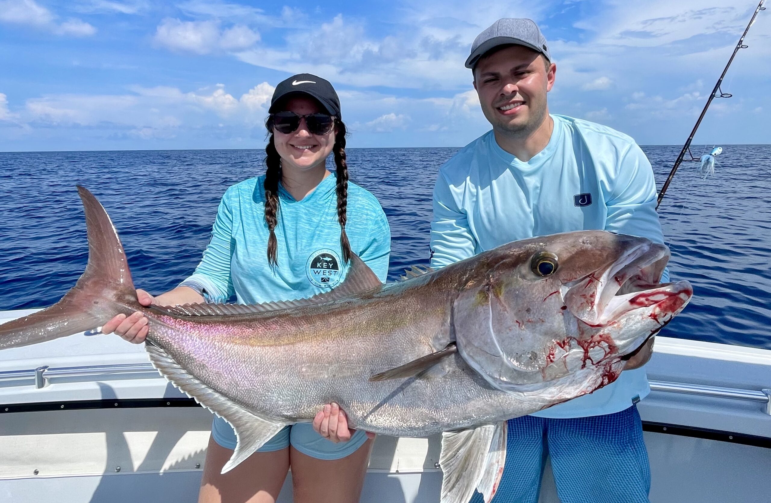 A couple on a fishing charter in Key Largo, Florida, proudly holding a massive Amberjack fish they caught. The background shows the beautiful waters of Key Largo.