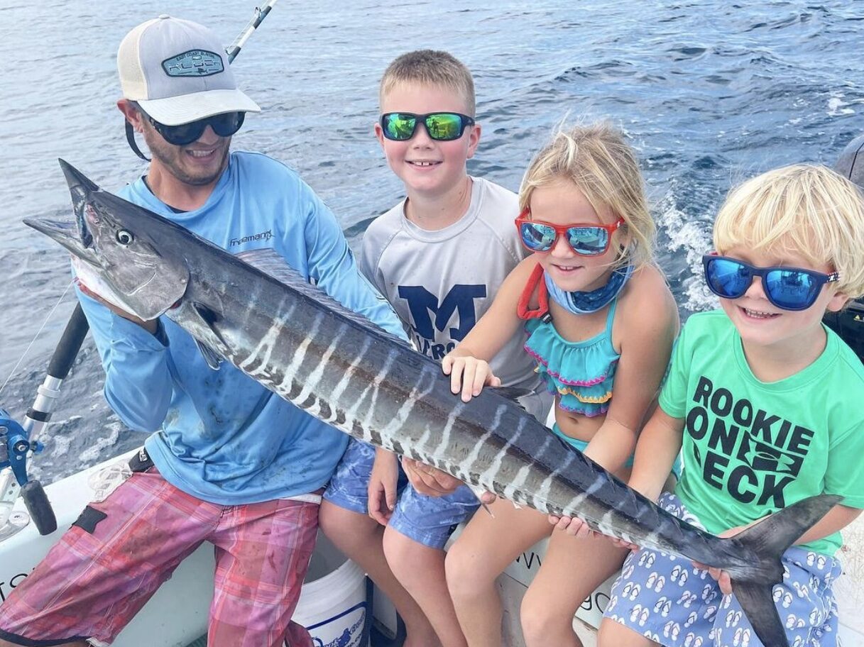 Three siblings on a family-friendly fishing charter during their vacation in Florida, proudly holding a wahoo fish they caught. The background showcases the beautiful ocean scenery, enhancing the excitement and joy of their fishing adventure.
