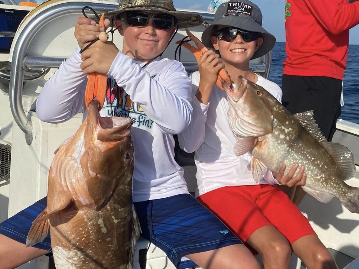 Two brothers holding two red groupers they caught during a fishing charter with their family. The background features the charter boat at the dock.