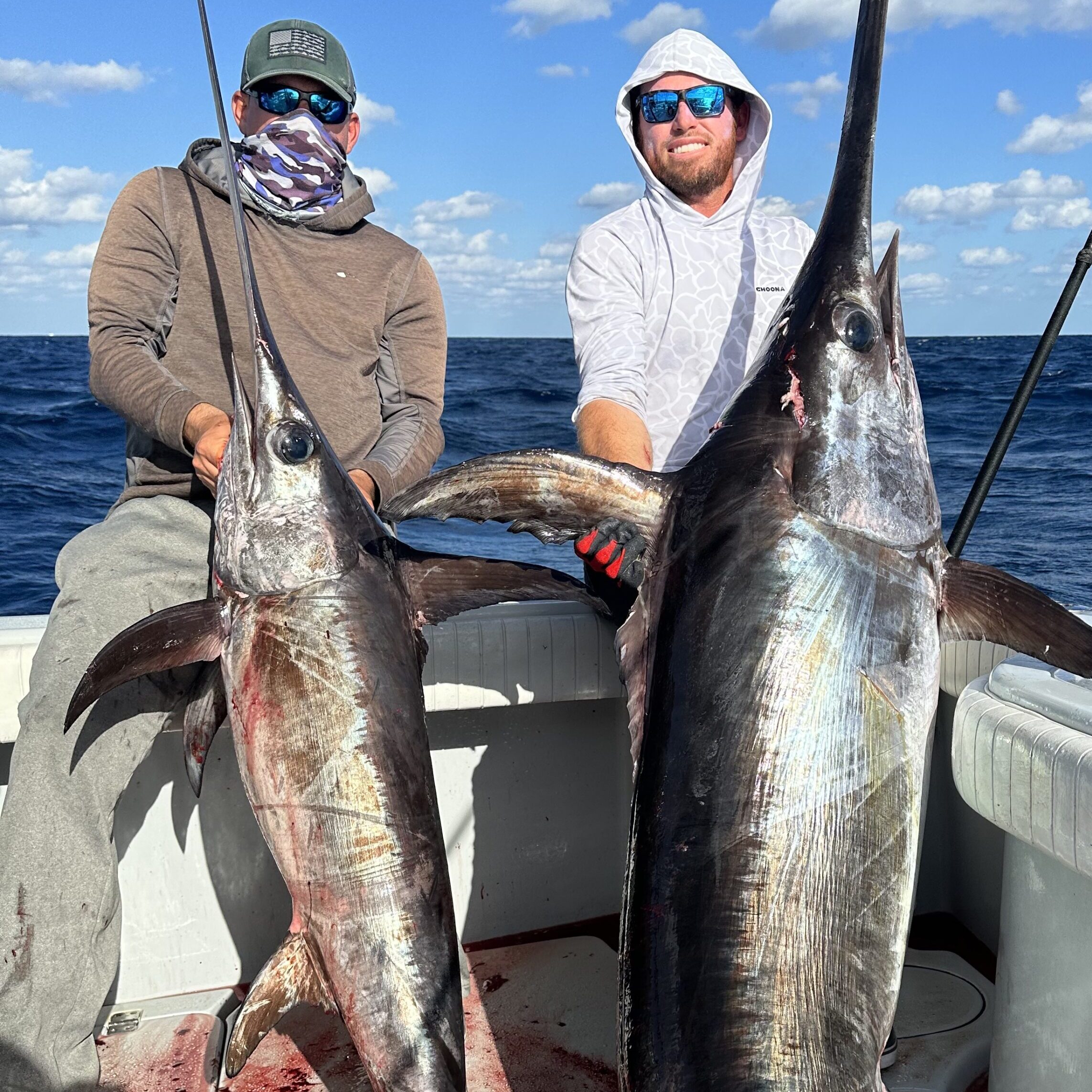 Two clients on a sword fishing charter proudly holding up two large swordfish they've caught, smiling with the ocean in the background.