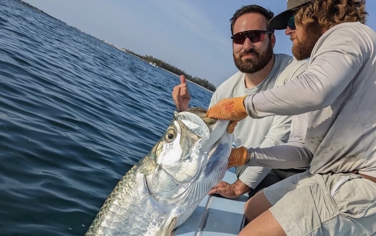 An angler proudly holds up a large tarpon, showcasing a successful fishing catch aboard a fishing charter. The tarpon's silvery scales glisten in the sunlight, and the background features a scenic view of the open sea with a clear blue sky and calm waters.