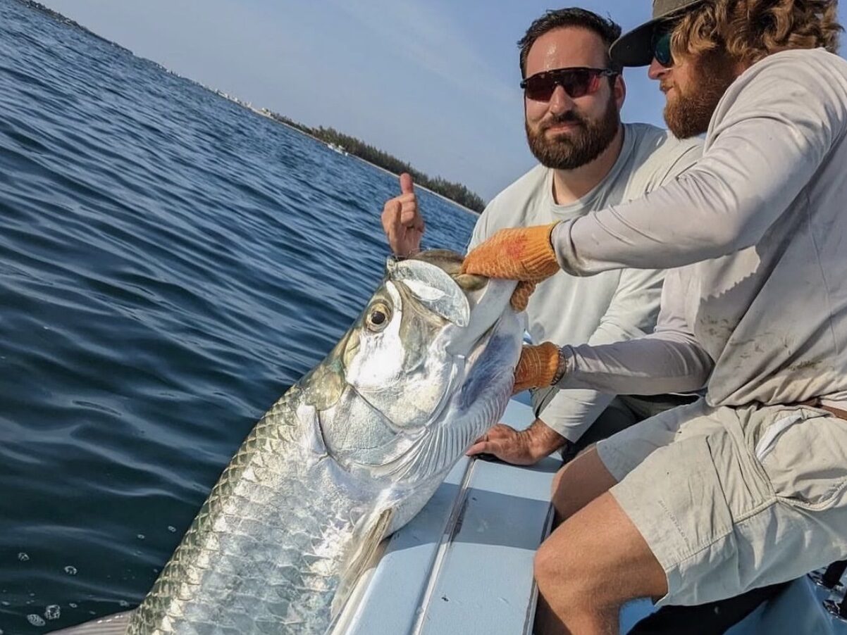Captain holds up a tarpon against the boat for a picture, with the client posing in the background, capturing a memorable moment during a tarpon fishing charter.