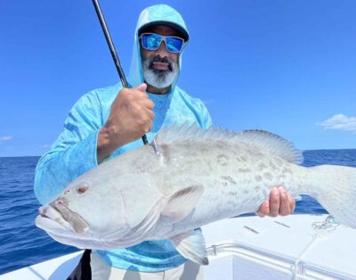 A man holding a snowy grouper caught during a fishing charter in Key West, Florida, with the ocean in the background.