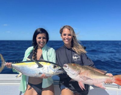 Two women proudly displaying a mutton snapper and tuna caught during a fishing charter on the ocean.