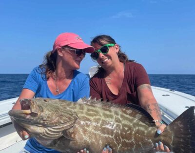 Two best friends showcasing a black grouper caught during a deep-sea fishing trip with their husbands.