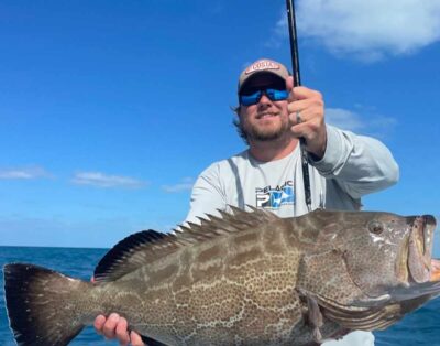 A man smiling for the camera while holding a big black grouper during a fishing charter with friends.